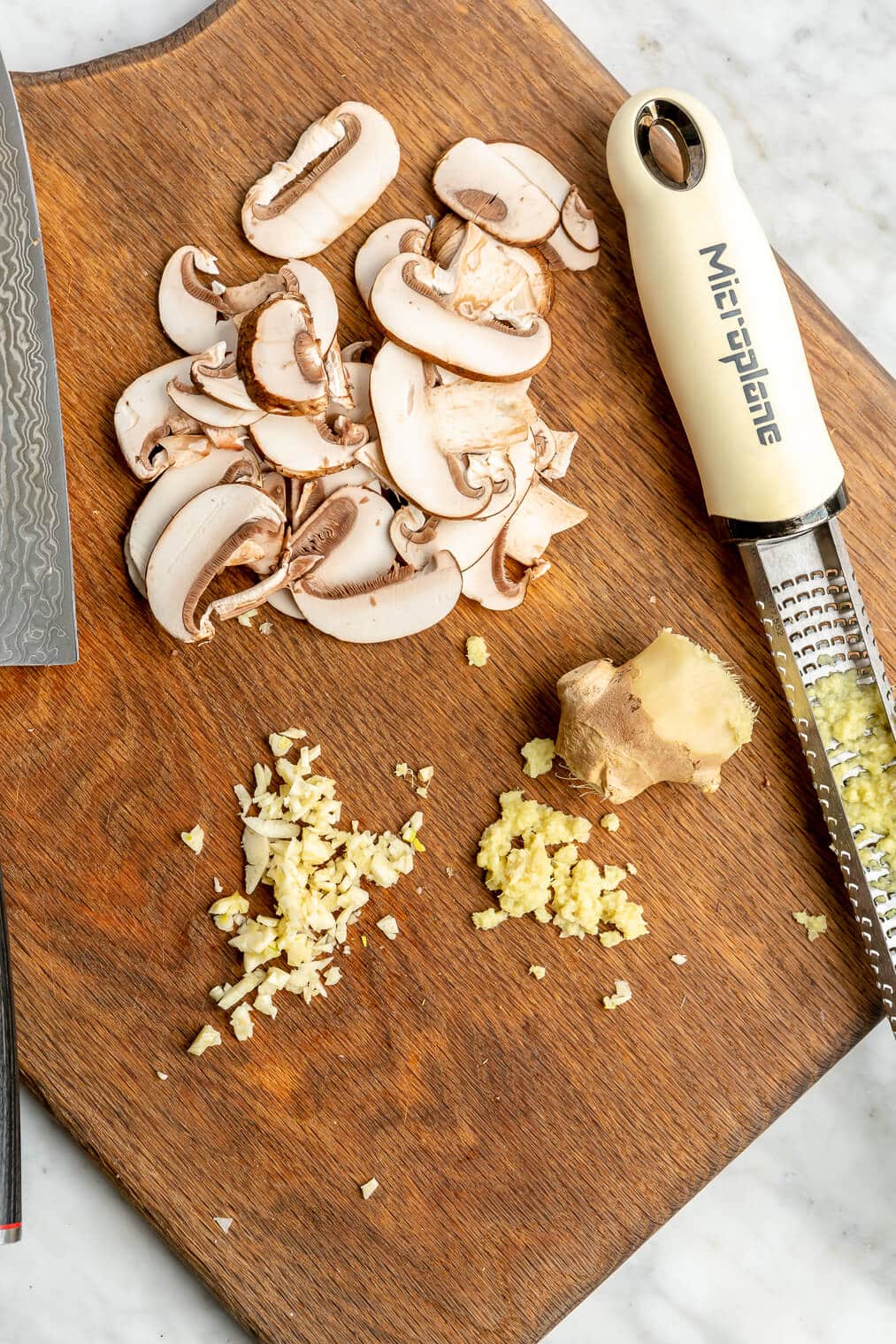 Sliced mushrooms, minced garlic, and grated ginger on a wooden cutting board.