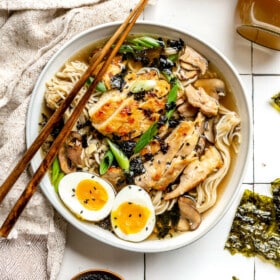 Bowl of chicken ramen on a white tile surface. The ramen is topped with a soft boiled egg, green onions, and black sesame seeds. There is a jar of bone broth to the right of the bowl and a small wooden bowl of black sesame seeds.