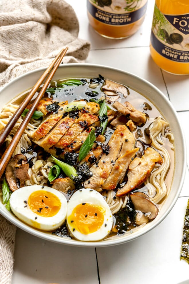 A bowl of chicken ramen on a white tile surface. The bowl is topped with green onions, a soft-boiled egg, and black sesame seeds.