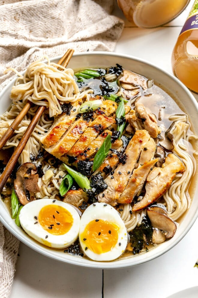 A bowl of chicken ramen on a white tile surface. The bowl is topped with green onions, a soft-boiled egg, and black sesame seeds. 