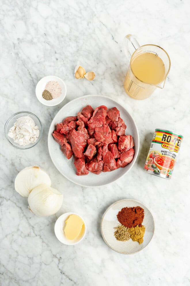 All of the ingredients for carne guisada - broth, garlic, salt, pepper, flour, onion, stew meat, Rotel, butter, and spices - sitting in small bowls on a marble surface.