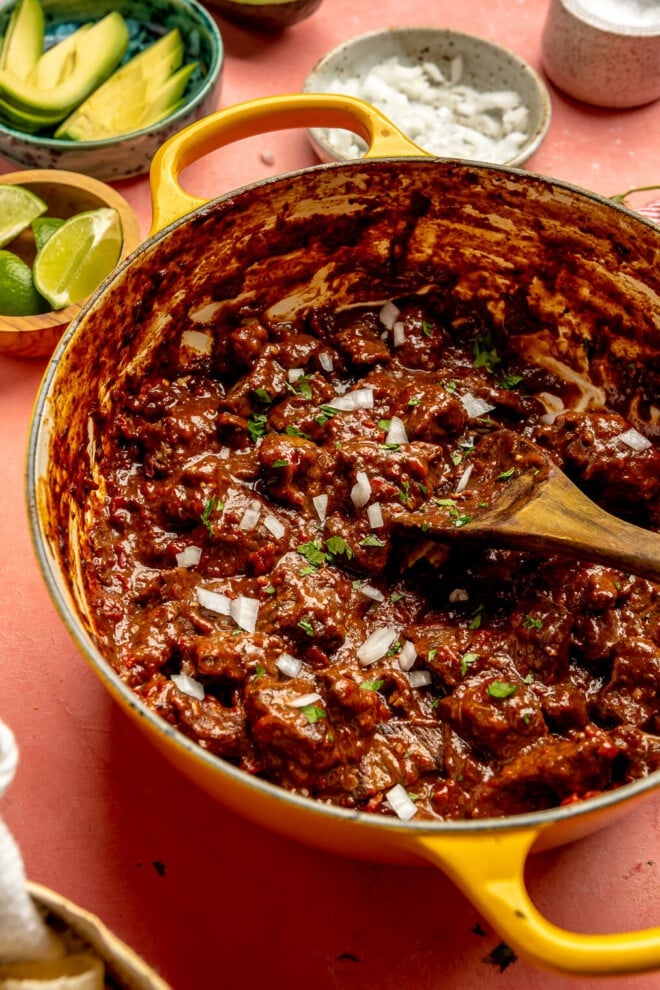 A large enameled cast iron pot filled with carne guisada topped with diced white onion and cilantro next to a bowl of sliced avocado, a bowl of lime wedges, and a bowl of diced white onion.