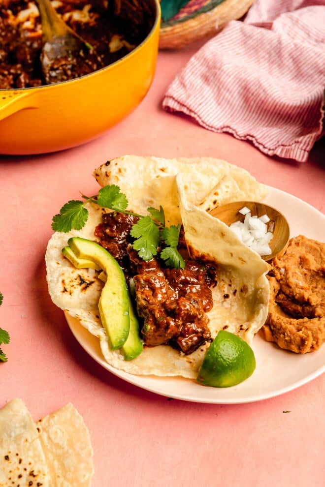 A carne guisada taco topped with cilantro and sliced avocado on a plate next to a serving of refried beans, a small bowl of diced white onion, and a lime wedge. 