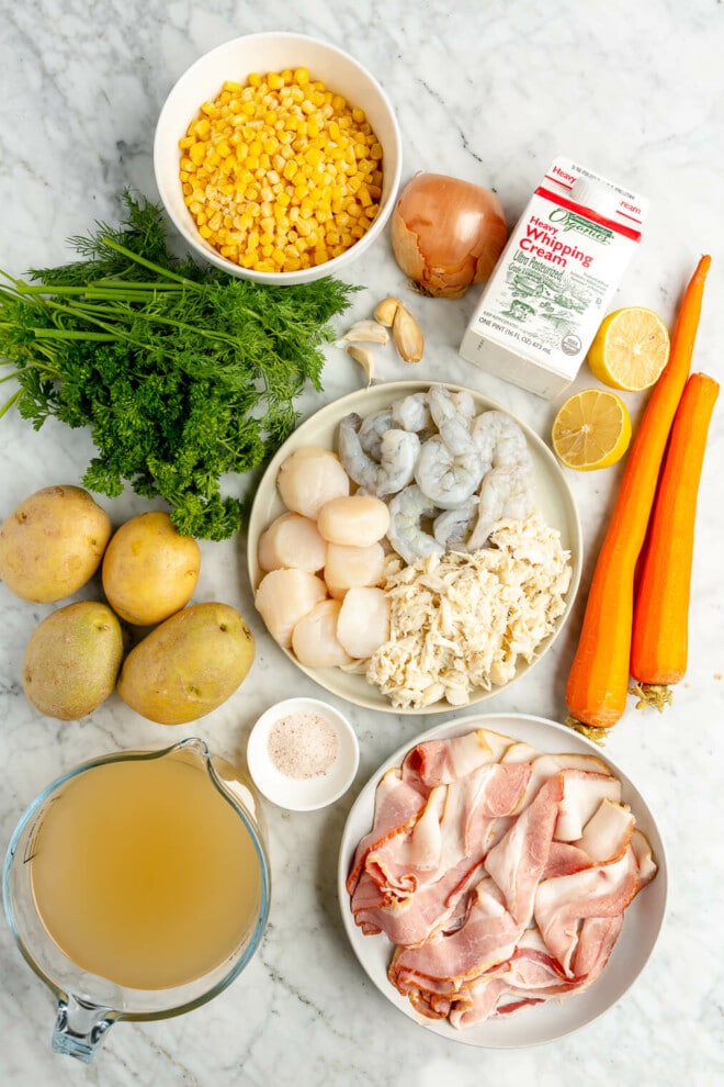 The ingredients for seafood chowder: broth, bacon, carrots, salt, crab meat, shrimp, scallops, potatoes, parsley, lemon, heavy cream, onion, garlic, and corn sitting on a marble surface.