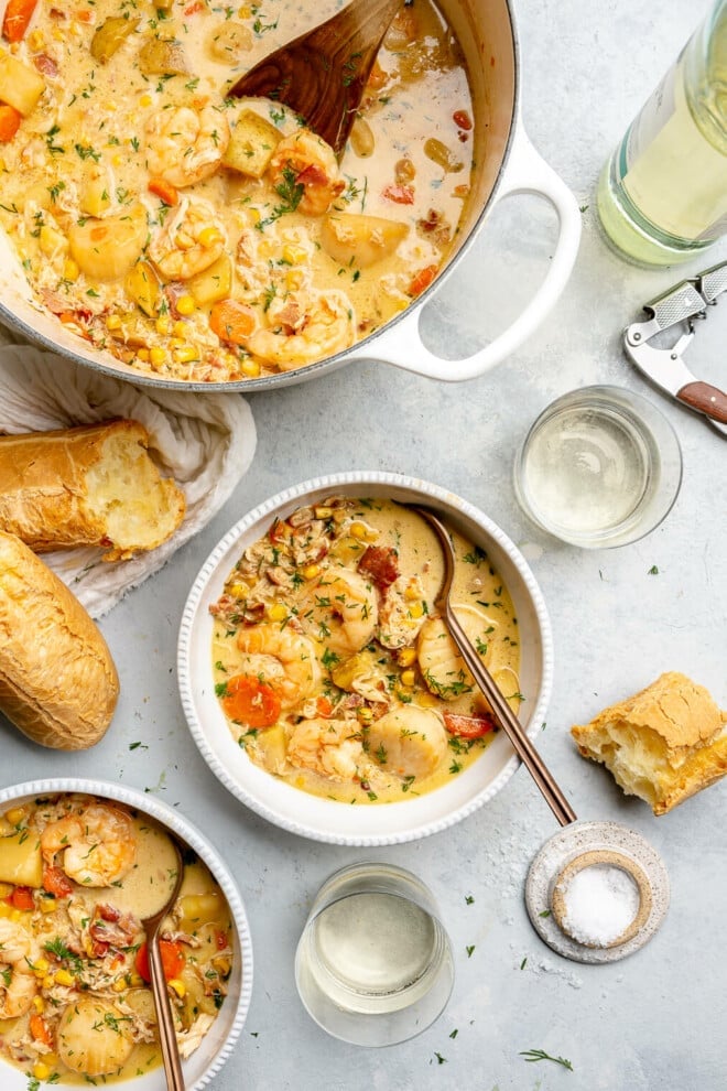 A single-serving bowl of seafood chowder next to another single-serving bowl of chowder, a large pot of seafood chowder, a couple of glasses of wine, and two baguettes. 