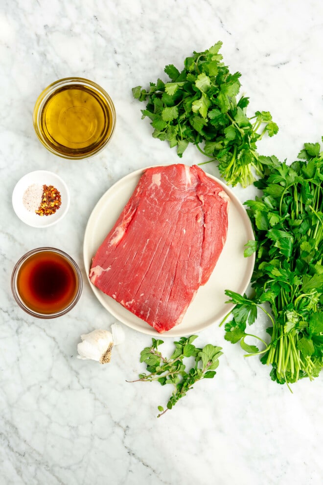 The ingredients for chimichurri flank steak: herbs, oil, vinegar, salt, red pepper flakes, garlic, and a flank steak all sitting on a marble surface