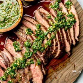 Sliced flank steak topped with chimichurri on a large wooden cutting board. Also pictured: a small bowl of chimichurri sauce and a small bowl of flaky sea salt