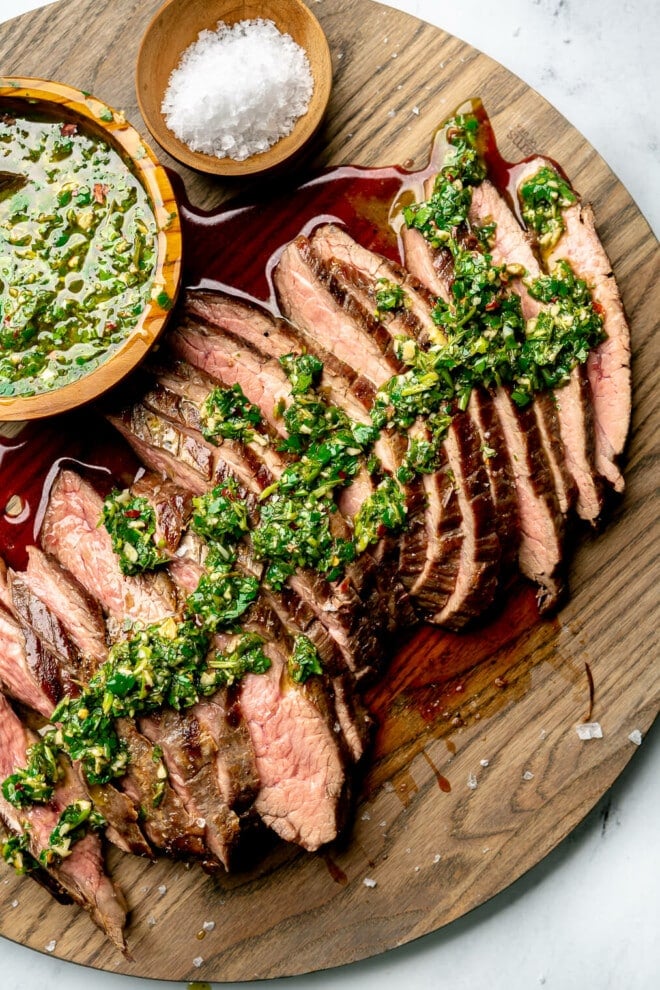 Sliced flank steak topped with chimichurri on a large wooden cutting board. Also pictured: a small bowl of chimichurri sauce and a small bowl of flaky sea salt