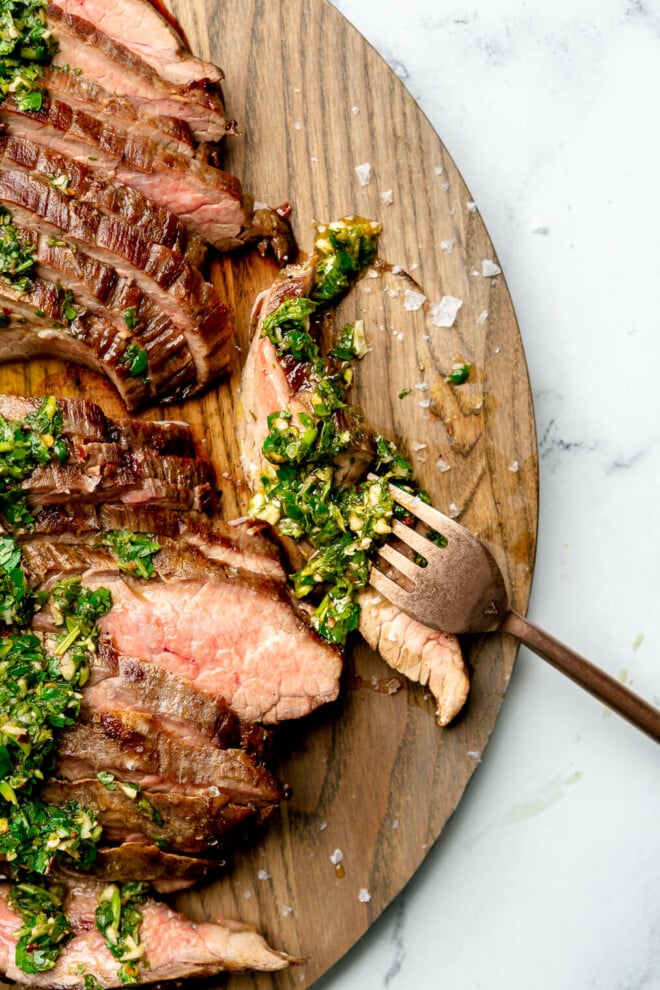 Sliced flank steak topped with chimichurri on a large wooden cutting board. Also pictured: a small bowl of chimichurri sauce and a small bowl of flaky sea salt