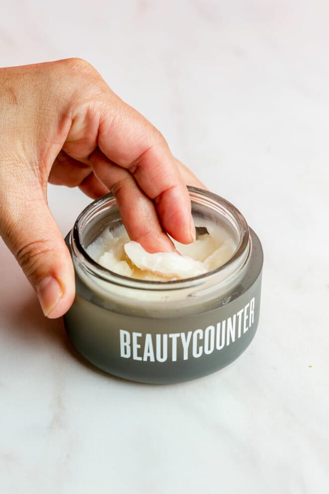 An open jar of Beautycounter's cleansing balm sitting on a marble surface and a person's finger getting some of the balm out of the jar.