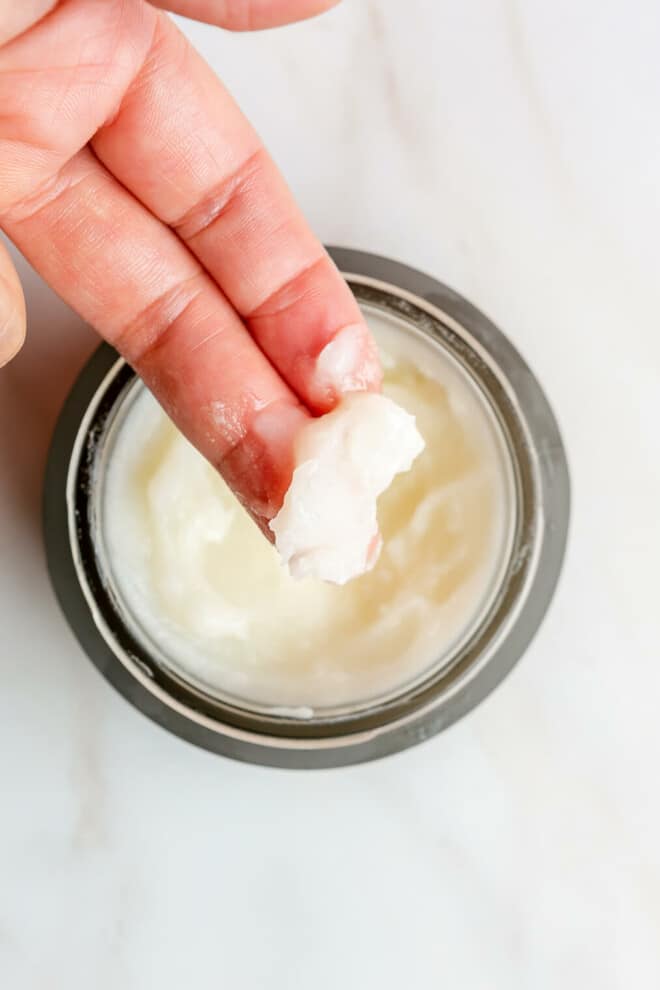 An open jar of Beautycounter's cleansing balm sitting on a marble surface and a person's finger getting some of the balm out of the jar.