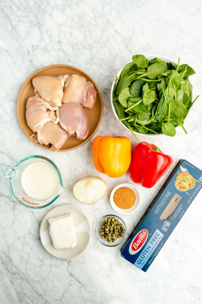 All of the ingredients for creamy cajun chicken pasta sitting on a marble surface. Chicken thighs, spinach, cream, cream cheese, bell peppers, onion, cajun seasoning, capers, and pasta.