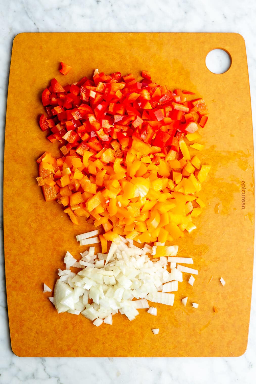 Diced red bell pepper, orange bell pepper, and white onion on an orange cutting board sitting on a marble surface.