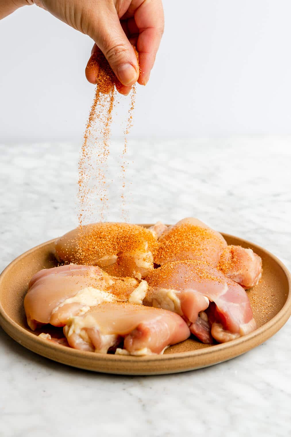 A person sprinkling cajun seasoning onto a plate of raw chicken thighs.