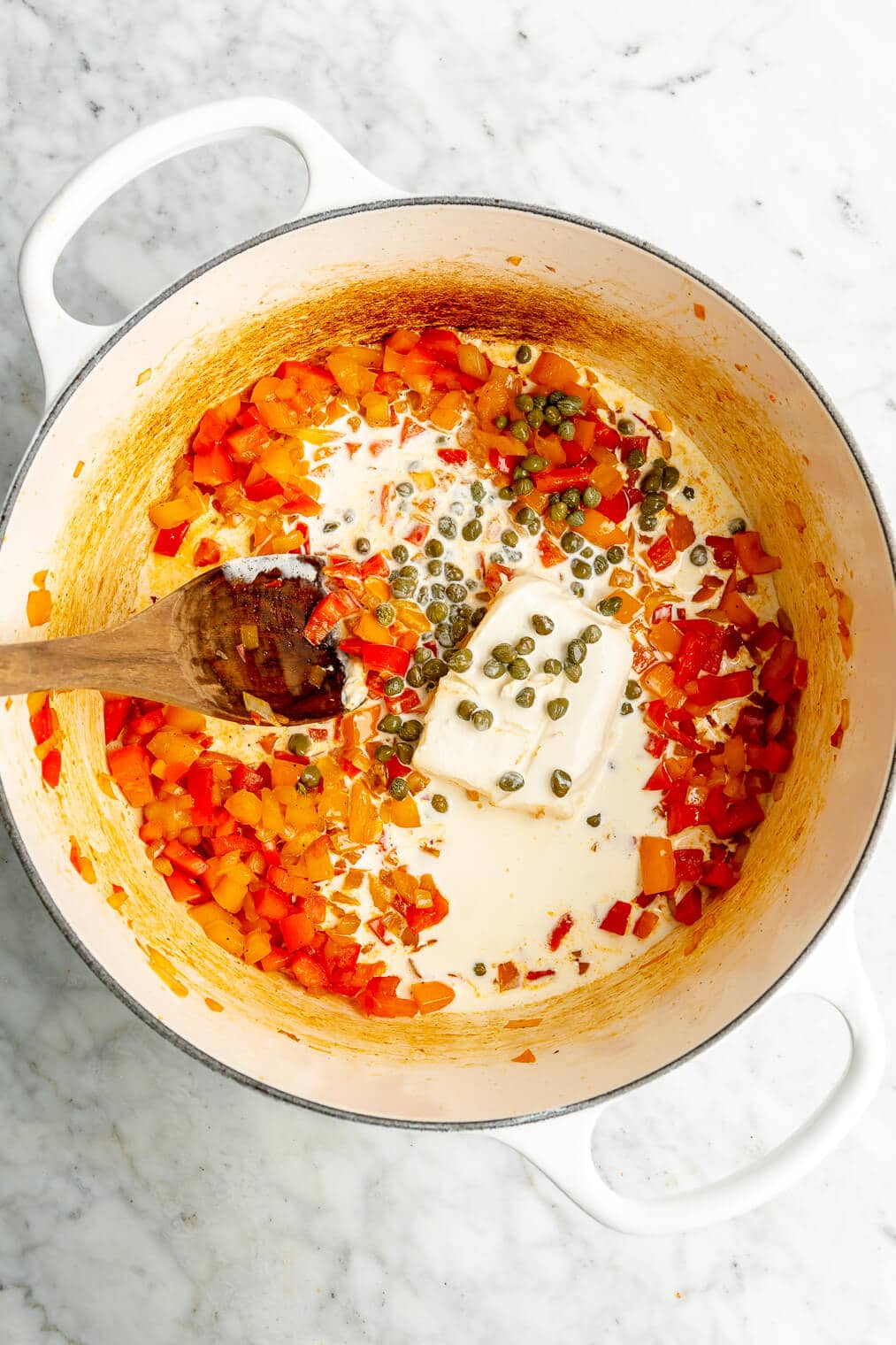 A top down view of sauteed bell peppers and onions, a block of cream cheese, heavy cream, and capers in an enameled cast iron pot before being stirred together to form a creamy cajun pasta sauce.