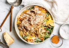 A large plate of creamy cajun pasta topped with cajun chicken thighs, lemon wedges, and grated parmesan cheese. Sitting next to the plate: a white kitchen towel, a small glass bowl of cajun seasoning, a bowl of flaky sea salt, a wedge of parmesan cheese, and two large serving spoons