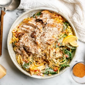 A large plate of creamy cajun pasta topped with cajun chicken thighs, lemon wedges, and grated parmesan cheese. Sitting next to the plate: a white kitchen towel, a small glass bowl of cajun seasoning, a bowl of flaky sea salt, a wedge of parmesan cheese, and two large serving spoons