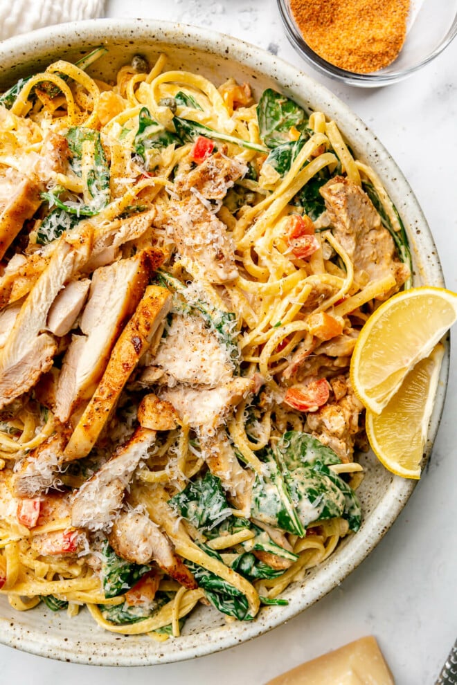 A large plate of creamy cajun pasta topped with cajun chicken thighs, lemon wedges, and grated parmesan cheese. Sitting next to the plate: a white kitchen towel, a small glass bowl of cajun seasoning, a wedge of parmesan cheese, and a microplane for grating the cheese