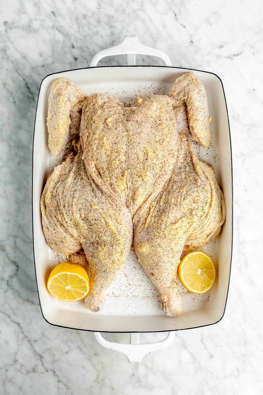A raw spatchcocked chicken rubbed with butter and seasoned with salt and pepper in a white casserole dish next to two lemon halves.
