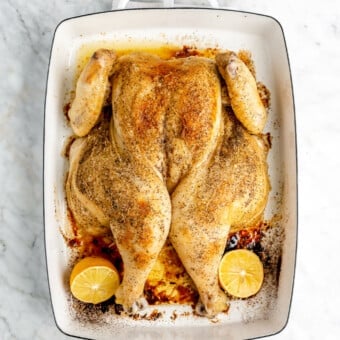 A cooked spatchcocked chicken rubbed with butter and seasoned with salt and pepper in a white casserole dish next to two lemon halves.