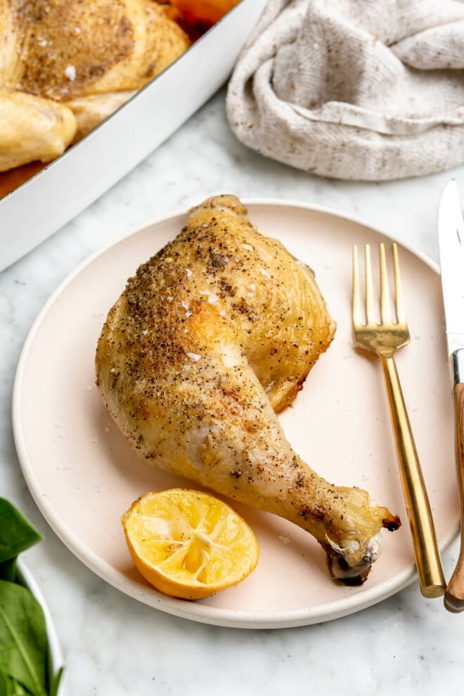 A cooked spatchcocked chicken rubbed with butter and seasoned with salt and pepper in a white casserole dish next to two lemon halves next to a plate with a chicken leg, lemon wedge, fork, and knife on it.