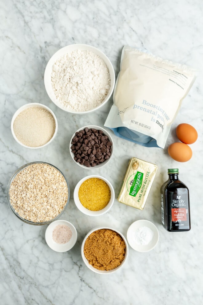 Ingredients for lactation cookies: butter, white sugar, brown sugar, two eggs, vanilla extract, collagen powder, brewer's yeast, cassava flour, quick oats, baking soda, salt, and chocolate chips sitting on a marble surface.