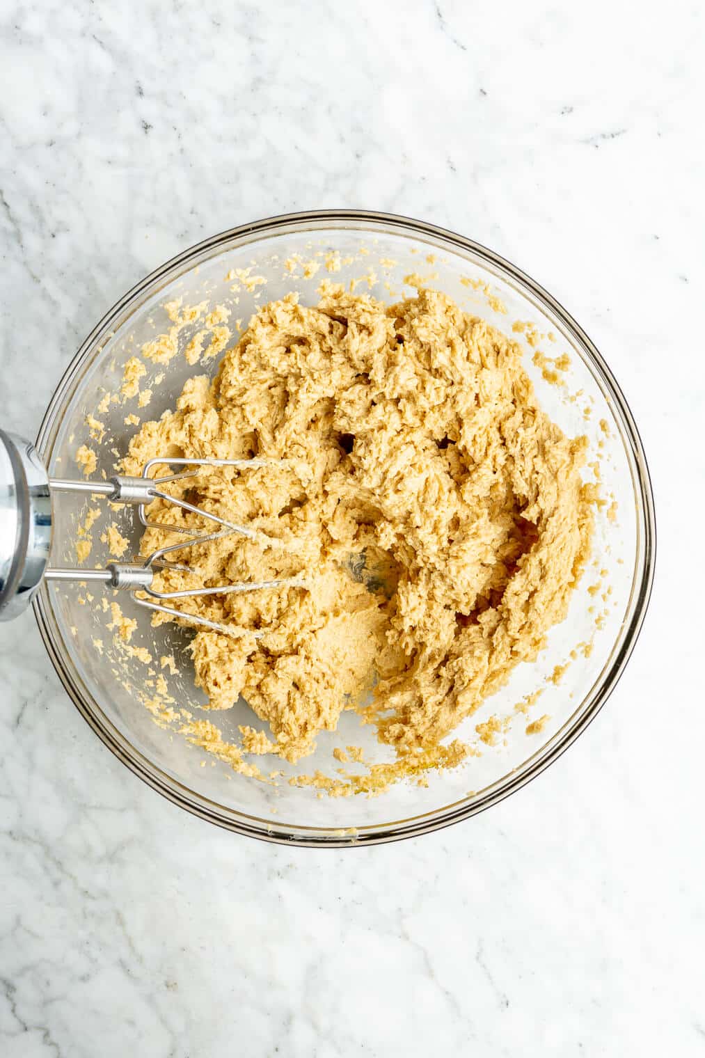 A large glass bowl of whipped softened butter, white sugar, brown sugar, eggs, and vanilla extract. Also in the photo: the whisks of the handheld electric mixer.