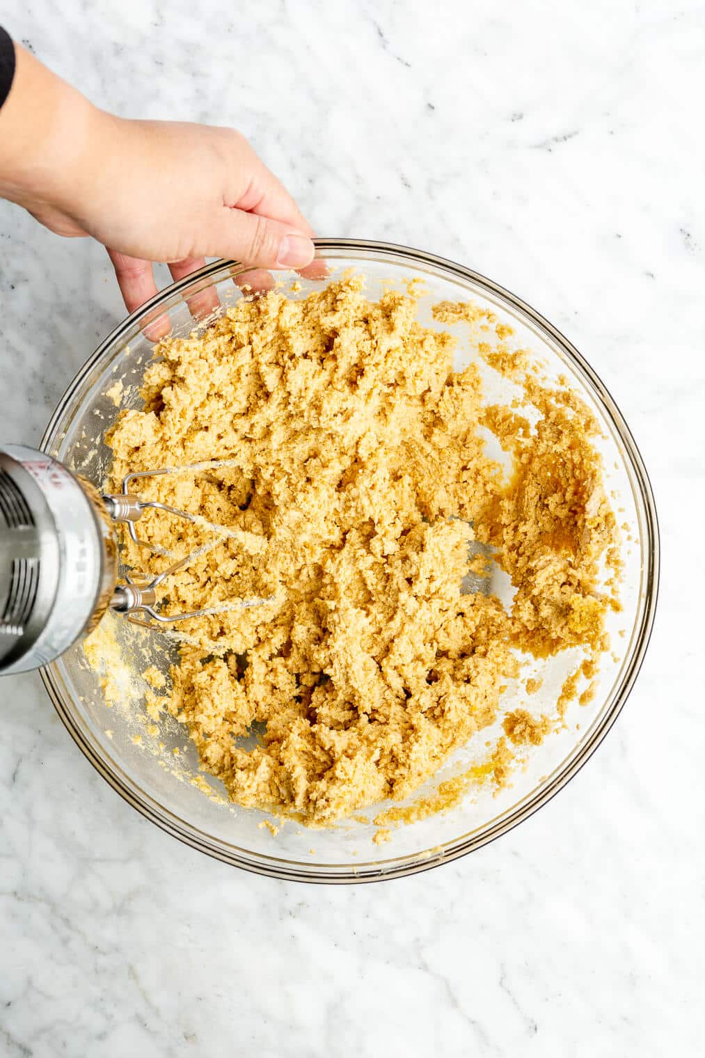 A person using an electric hand mixer to beat a cookie dough mixture in a large glass bowl.