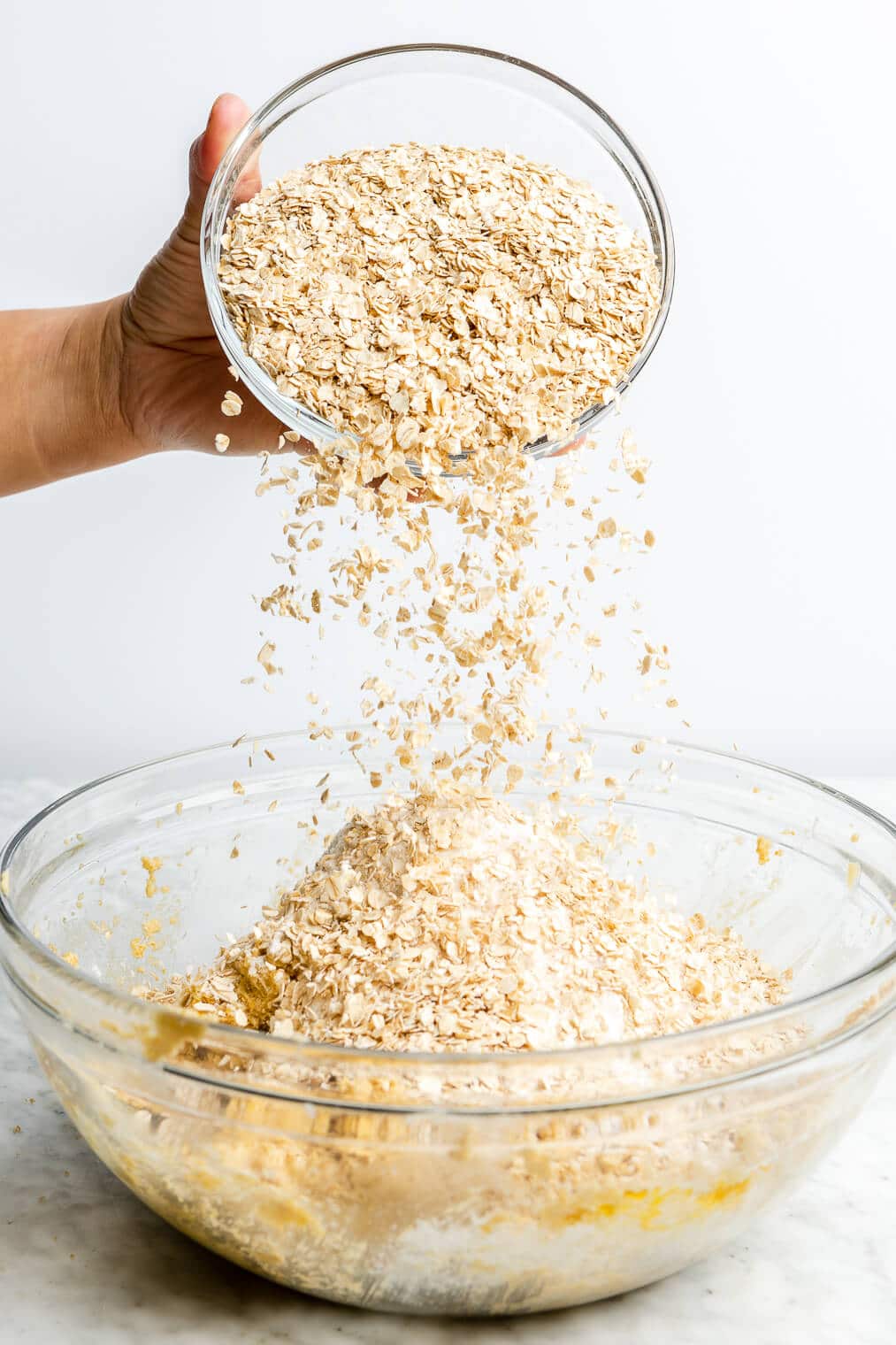 Two cups of quick oats being added to a large bowl of cookie dough mixture.