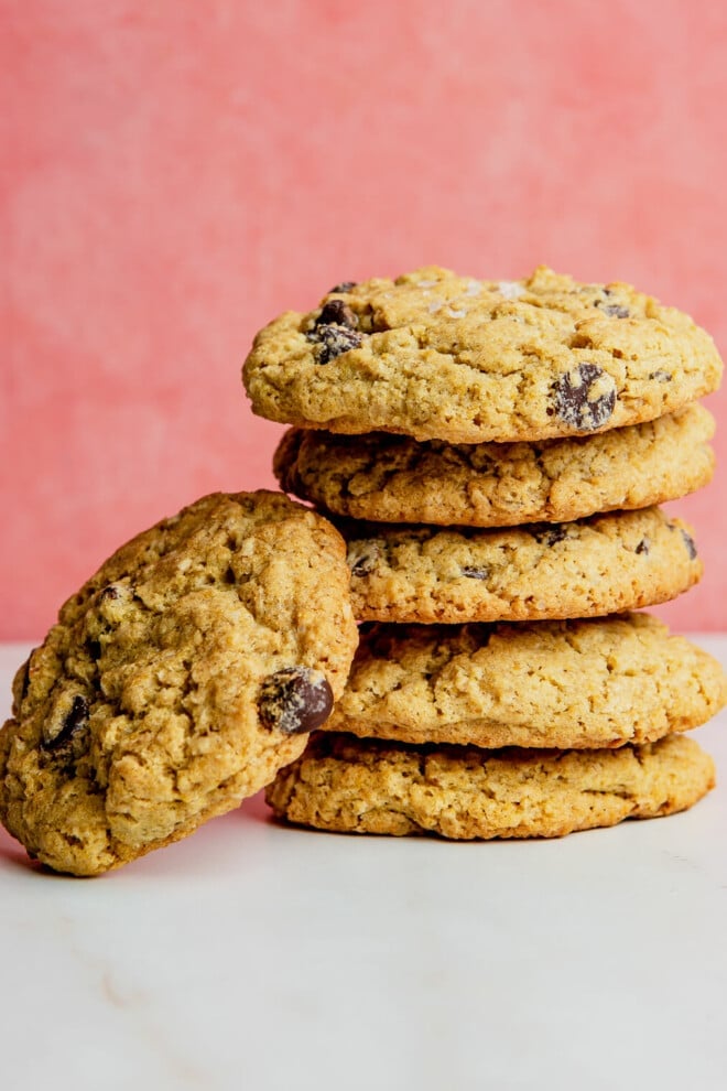 A stack of 6 oatmeal chocolate chip lactation cookies on a marble surface with a pink background.