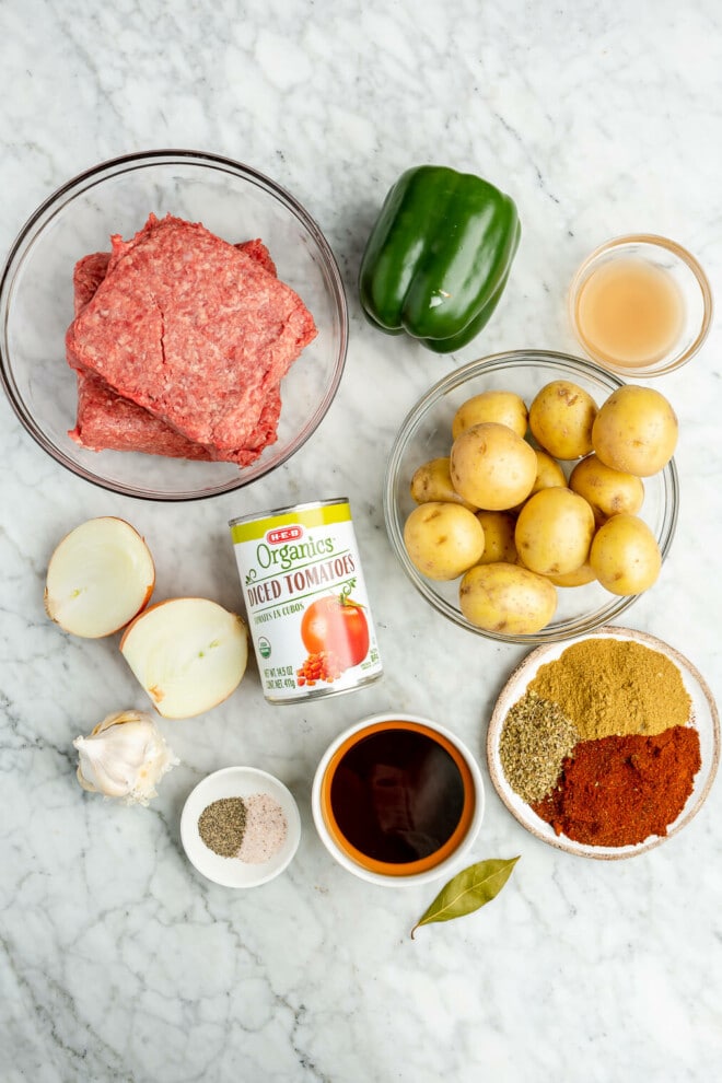 Ingredients for beef picadillo: a bowl of raw ground beef, a green bell pepper, apple cider vinegar, Yukon gold potatoes, a can of diced tomatoes, yellow onion, a bulb of garlic, salt and pepper, beef broth, chili powder, cumin, oregano, and a bay leaf all sitting on a marble surface