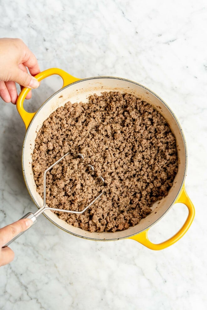 A person's hands using a metal potato masher to cook and crumble ground beef in an enameled cast iron pot.