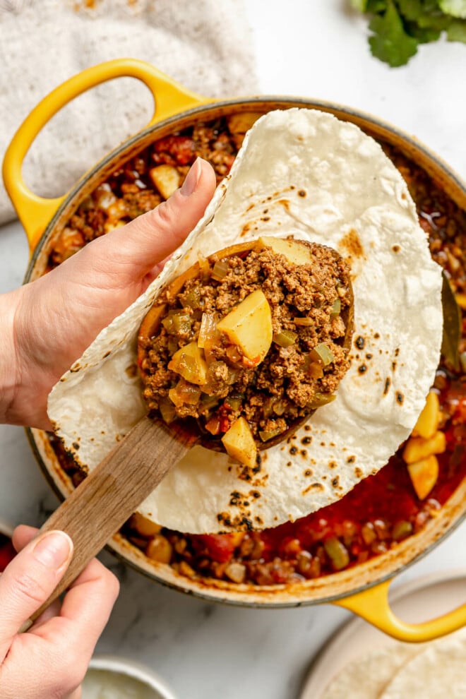 Picadillo being spooned into a charred flour tortilla held over the pot of picadillo