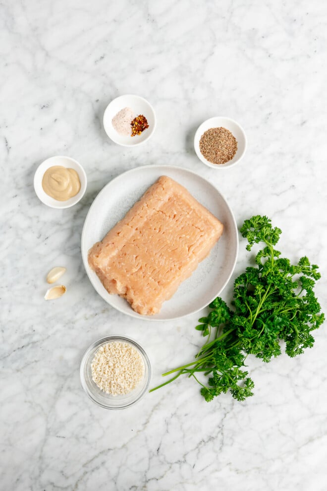 The ingredients for homemade chicken burgers (raw ground chicken, panko breadcrumbs,parsley, garlic, dijon mustard, flaxseed, salt, and red pepper flakes) on a marble surface.