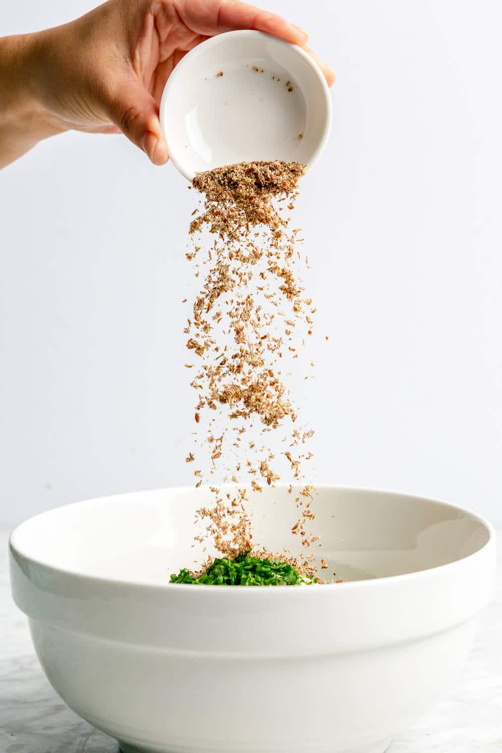 A small measuring cup of flax meal being added to a large bowl of ground chicken, fresh parsley, and panko bread crumbs to make homemade chicken burgers.