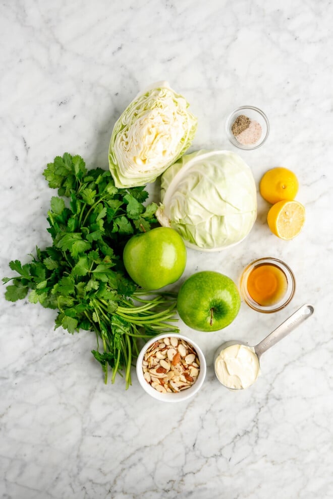 All of the ingredients for green apple slaw (cilantro, almonds, mayo, honey, lemon, green apple, green cabbage, salt, and pepper) on a marble surface.