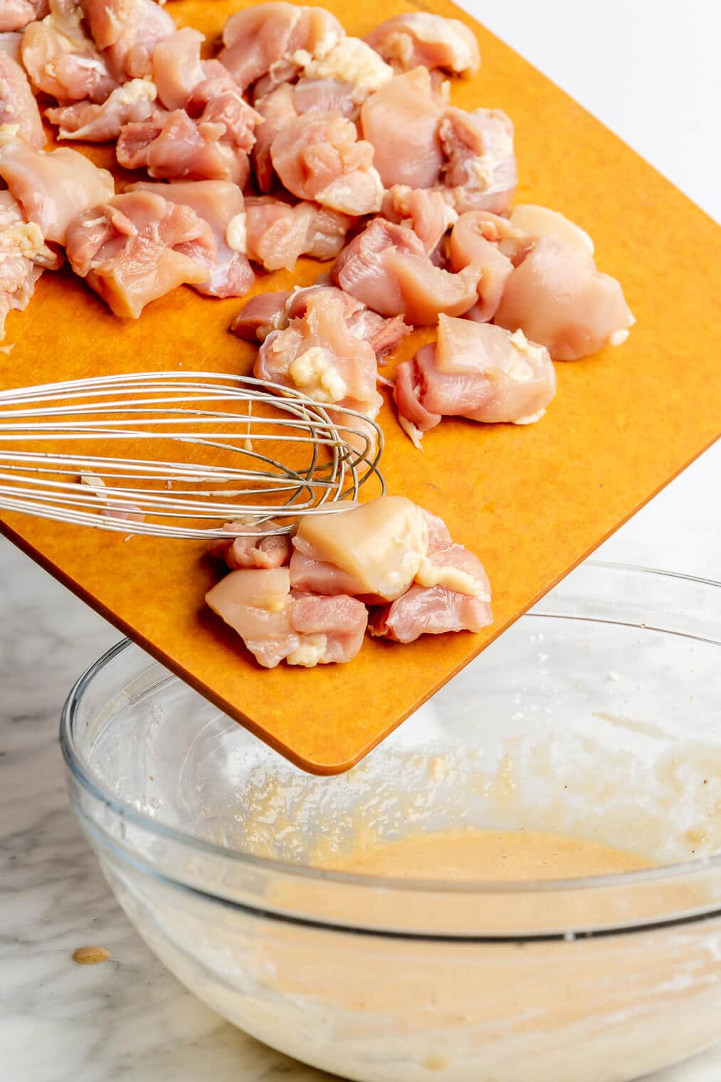 A person using a small whisk to guide chopped chicken thighs into a large glass bowl of whisked egg white, arrowroot starch, coconut aminos, apple cider vinegar, and salt.