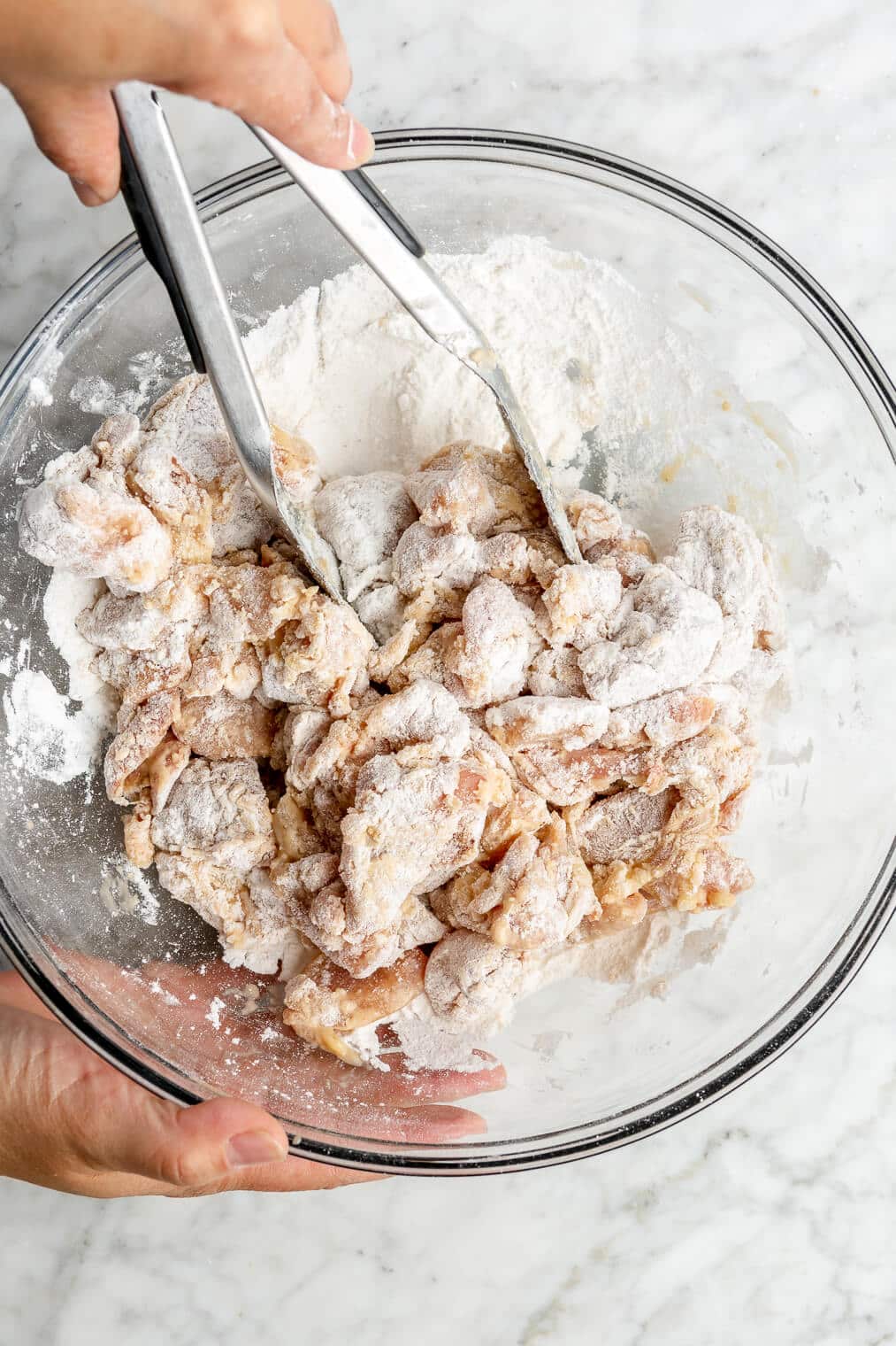 A person using tongs to coat small chunks of raw chicken thighs to a large glass mixing bowl of arrowroot starch, white rice flour, and salt.