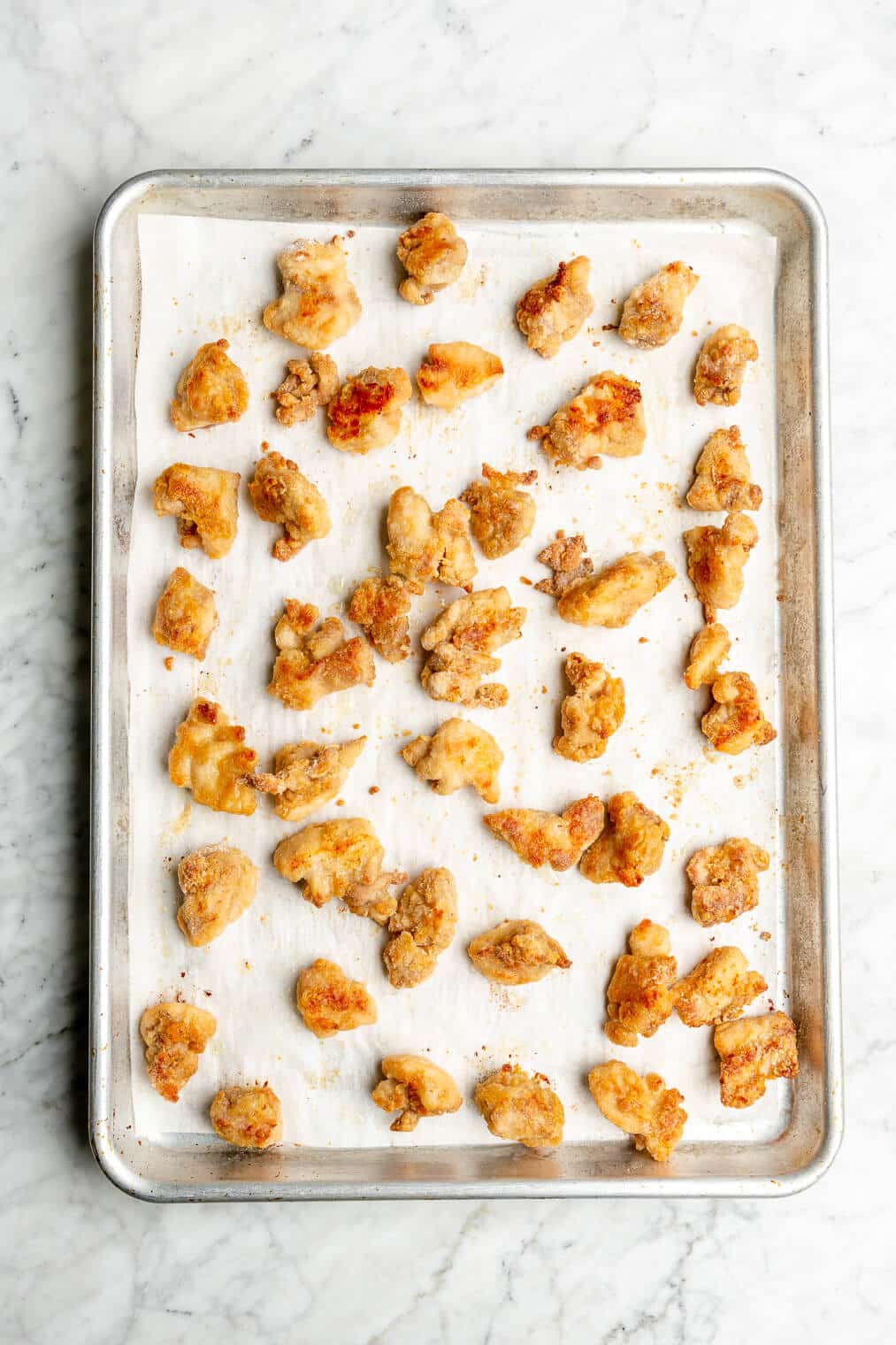 Small chunks of crispy chicken thighs coated with an arrowroot starch and white rice flour coating spread out on a parchment paper lined baking sheet.