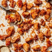 A parchment paper lined sheet pan with honey sesame chicken on it. Also on the sheet pan: a jar of honey sesame sauce, a small wooden bowl of sesame seeds, and two spoons.