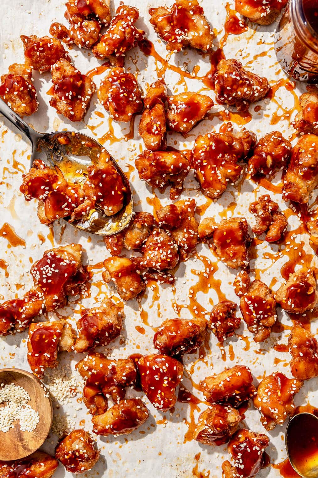 A parchment paper lined sheet pan with honey sesame chicken on it. Also on the sheet pan: a jar of honey sesame sauce, a small wooden bowl of sesame seeds, and two spoons.