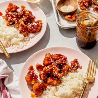 Two light pink plates, each with a portion of honey sesame chicken and white rice. Also pictured: a parchment paper lined sheet pan of honey sesame chicken and a glass of ice water.