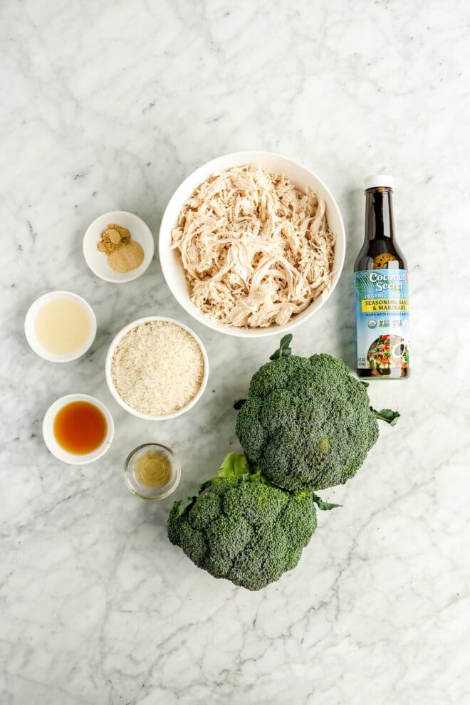 All of the ingredients for teriyaki chicken bowls (shredded chicken, coconut aminos, two heads of broccoli, dry rice, spices, vinegar, sesame oil, and fish sauce) on a marble surface.