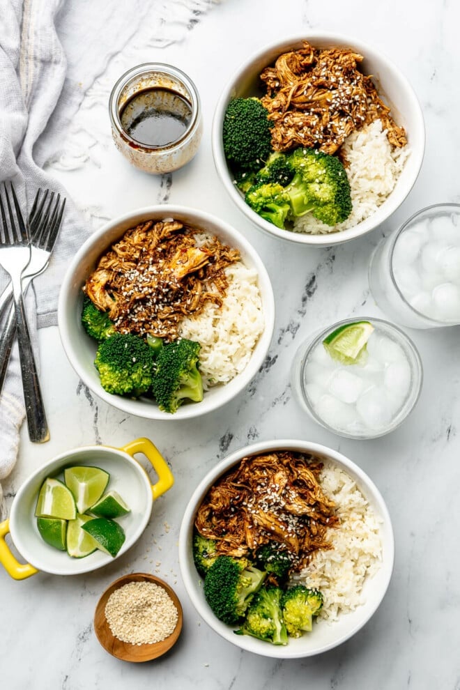 Three teriyaki chicken bowls on a marble surface next to two glasses of ice water, a bowl of quartered limes, a small bowl of sesame seeds, and a jar of homemade teriyaki sauce.