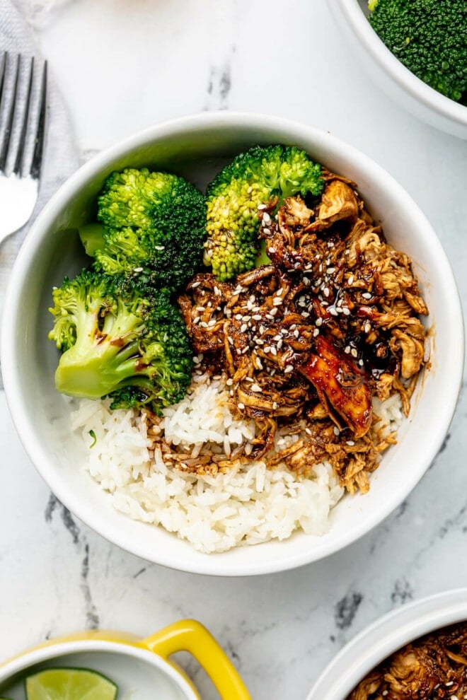 Three teriyaki chicken bowls on a marble surface next to two glasses of ice water, a bowl of quartered limes, a small bowl of sesame seeds, and a jar of homemade teriyaki sauce.