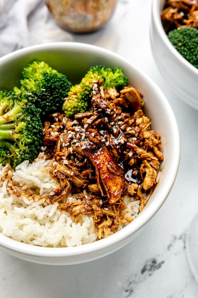 Three teriyaki chicken bowls on a marble surface next to two glasses of ice water, a bowl of quartered limes, a small bowl of sesame seeds, and a jar of homemade teriyaki sauce.