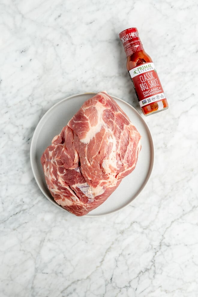 A large raw pork butt on a plate next to a bottle of Primal Kitchen Classic BBQ Sauce on a marble surface.