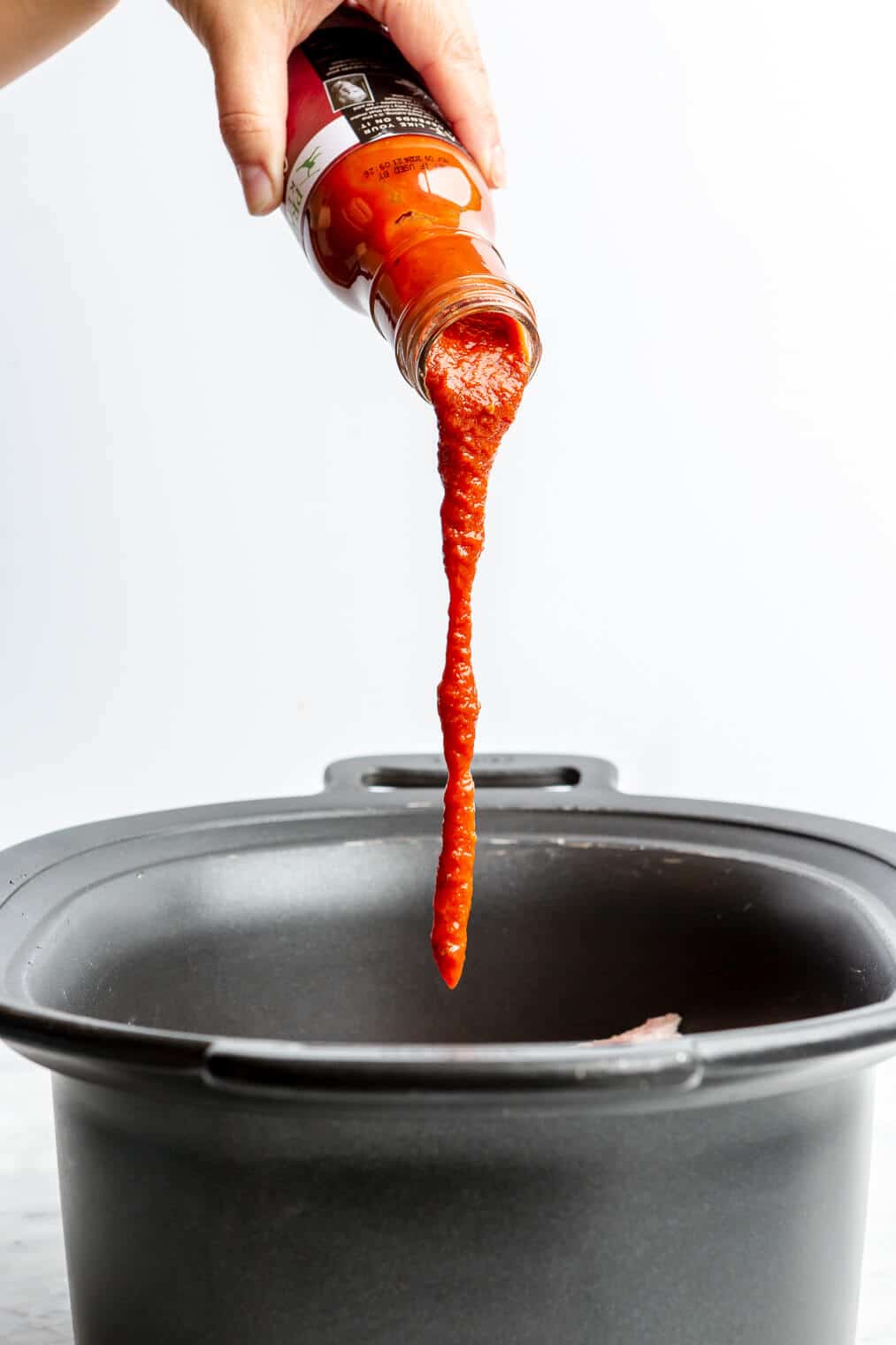 A jar of BBQ sauce being poured into a large crock pot of raw pork butt.