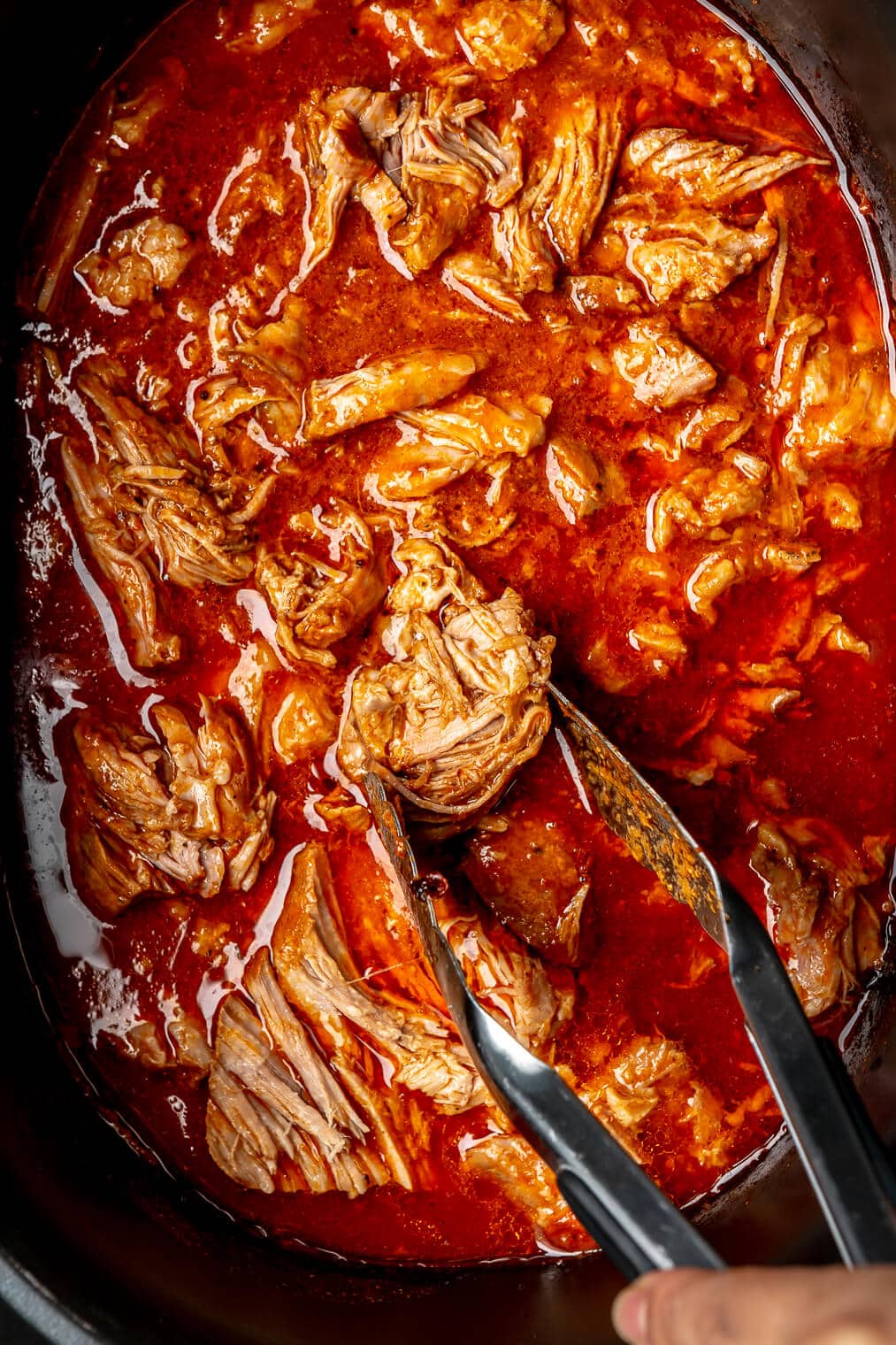 A crock pot of BBQ pulled pork. Some of the pork is being pulled out with metal tongs.