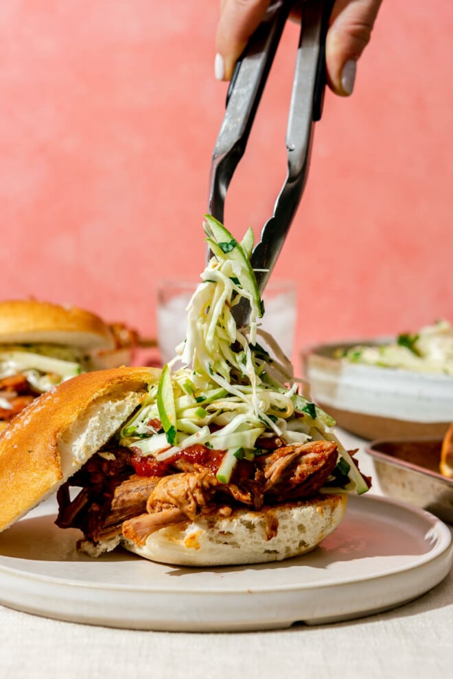 A person using metal tongs to place green apple slaw on top of a pulled pork sandwich.